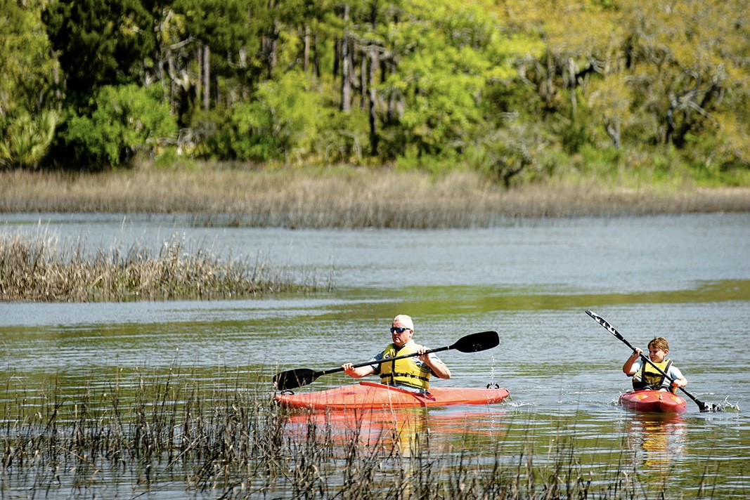 Father and son kayaking through the marsh grass exploring the tidal creeks of Palmetto Bluff in red kayaks.