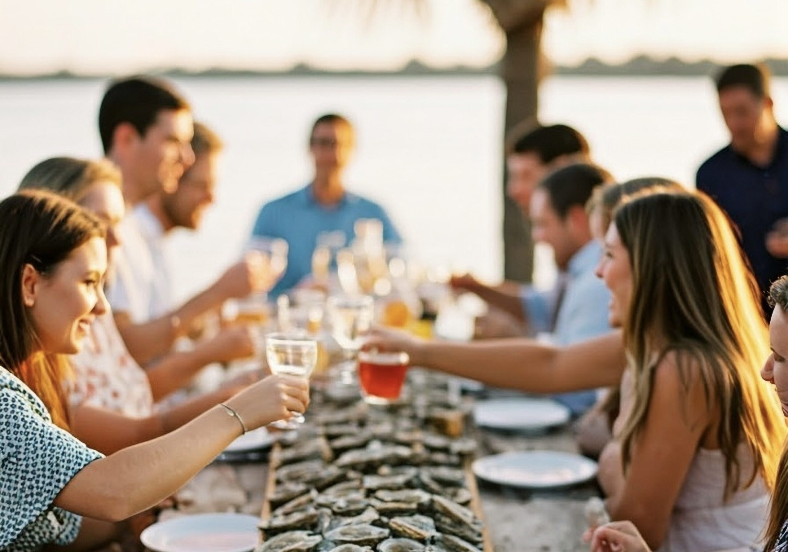 friends around oysters at an outdoor table setting with water in background