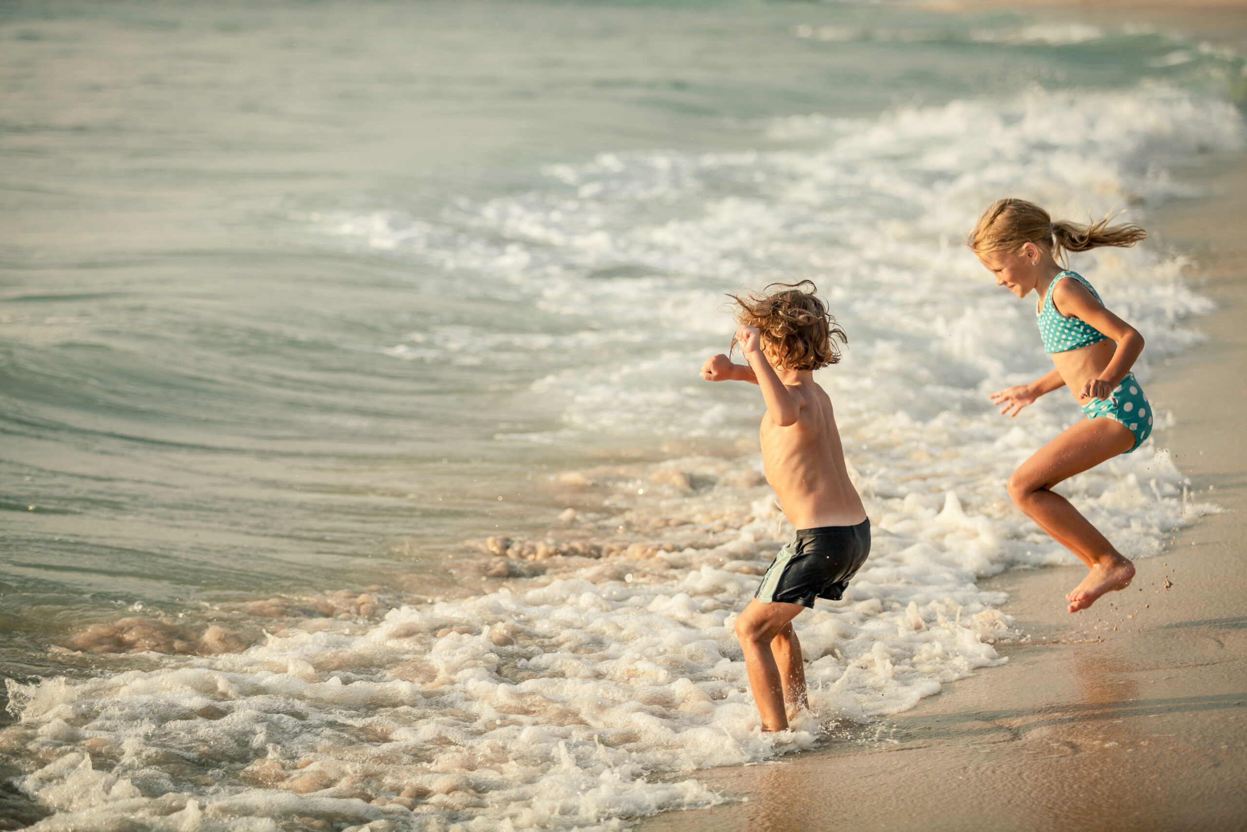 a boy and a girl jumping in the water at the beach on a sunny day